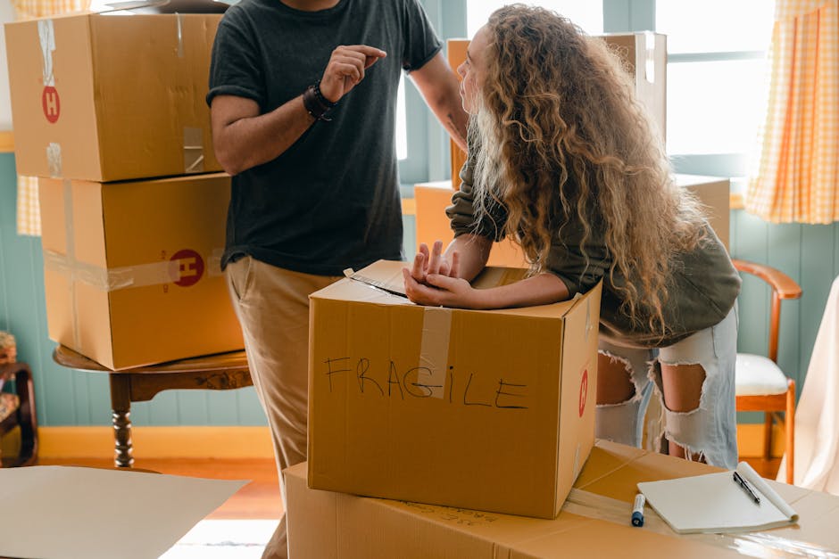 A person with a black shirt and tan trousers is assisting a woman with long, curly blonde hair and torn jeans, who is leaning over a medium-sized cardboard box labeled 'FRAGILE'. The box is positioned on a table inside a bright, well-lit room with blue walls, wooden flooring, and orange-checkered curtains. In the background, there are large cardboard boxes stacked on a wooden side table, some wrapped with clear plastic tape, indicating packing for a house relocation. On the table near the cardboard box, there are a white notepad and a black marker, suggesting an organized packing process by [COMPANY_NAME]. The scene depicts the careful handling and packing of household items, an essential part of professional removals and furniture transport services offered by [COMPANY_NAME], to ensure safe and efficient home moving.