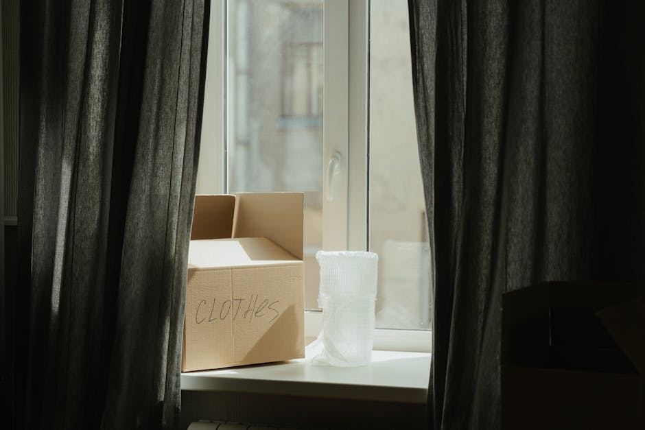 A cardboard box labeled 'clothes' placed on a white windowsill inside a room, partially covered by dark curtains framing the window. The box contains clothing and is positioned next to a roll of white packing paper or fabric. Outside the window, an outdoor building wall is visible. The scene suggests an ongoing home relocation or packing process, with furniture and packing materials prepared for transport. The interior environment is well-lit by natural daylight, highlighting the packing materials and the window's view, as part of the moving process facilitated by Removals House, a professional removal service specializing in furniture transport and packing during house moves.