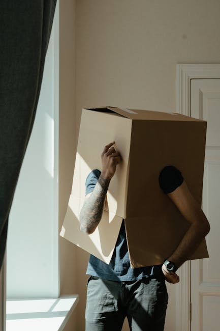 A person with a black shirt and tan trousers is assisting a woman with long, curly blonde hair and torn jeans, who is leaning over a medium-sized cardboard box labeled 'FRAGILE'. The box is positioned on a table inside a bright, well-lit room with blue walls, wooden flooring, and orange-checkered curtains. In the background, there are large cardboard boxes stacked on a wooden side table, some wrapped with clear plastic tape, indicating packing for a house relocation. On the table near the cardboard box, there are a white notepad and a black marker, suggesting an organized packing process by [COMPANY_NAME]. The scene depicts the careful handling and packing of household items, an essential part of professional removals and furniture transport services offered by [COMPANY_NAME], to ensure safe and efficient home moving.