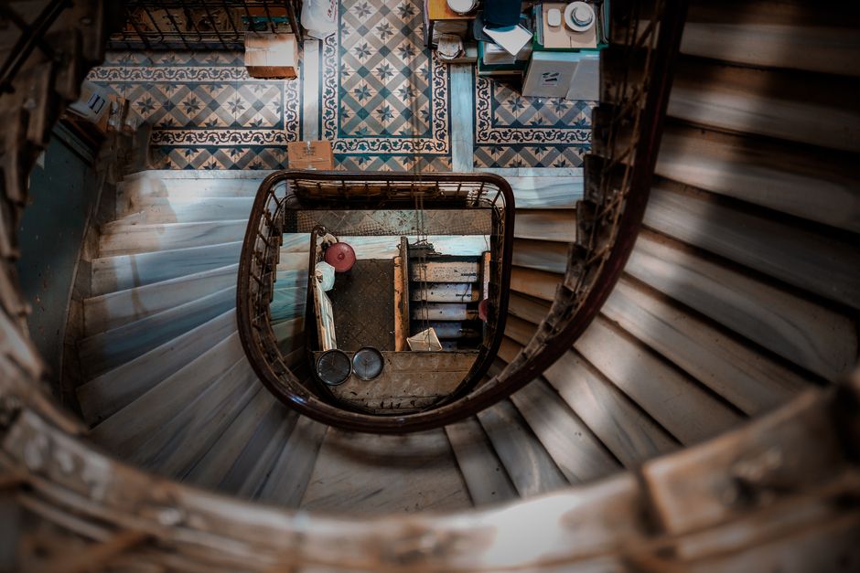 View looking down the central spiral staircase of a residential property, with wooden handrails and white marble steps arranged in a circular pattern. At the bottom of the staircase, a small landing area is visible, featuring various packing materials, including cardboard boxes, bubble wrap, and tissue paper, along with some household items such as a pink ball, metal bowls, and a wooden box. The interior space is lit by natural light, revealing decorative patterned flooring tiles and a doorway leading to an adjacent room filled with additional packing supplies and boxes. This image illustrates the preparatory stages of a home relocation, emphasizing the importance of careful packing and organized furniture transport, as performed or supervised by a professional removal service like Removals House during the process of moving fragile antiques or other valuables across the UK.
