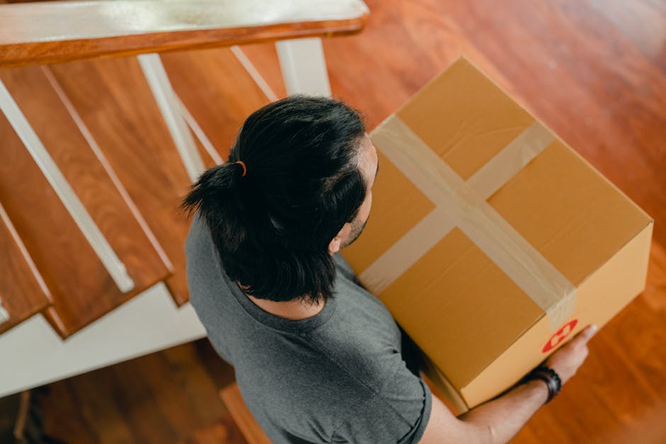 Overhead view of a person with dark hair tied back in a ponytail, wearing a grey t-shirt, holding a medium-sized cardboard moving box sealed with packing tape in both hands. The box has a red recycling symbol on one side. The individual is standing in a room with wooden flooring, adjacent to a staircase with white banisters. The scene depicts the process of packing or unpacking during a home relocation, with the person preparing to load or unload the box. The environment is well-lit, possibly by natural light, with clear visibility of the packing materials and furniture nearby. This visual relates to furniture transport and packing and moving activities carried out by Removals House as part of their house relocation services.